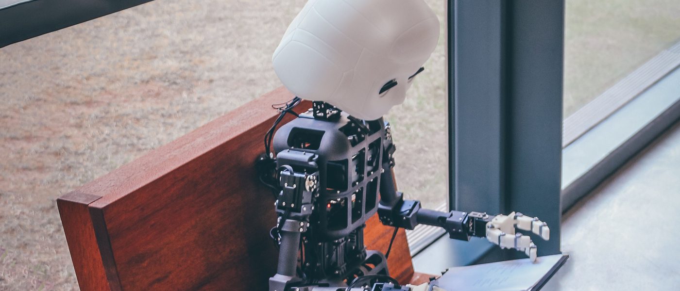 A humanoid robot sitting on a bench reading from a laptop